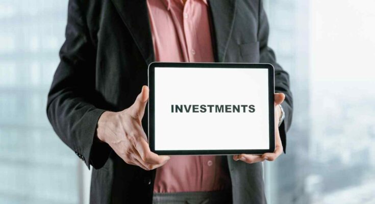 A business professional in a dark suit and red shirt hold tablet display the word ''investment'' against a blurred office style background